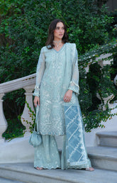 Woman in a light blue embroidered outfit standing outdoors with greenery in the background