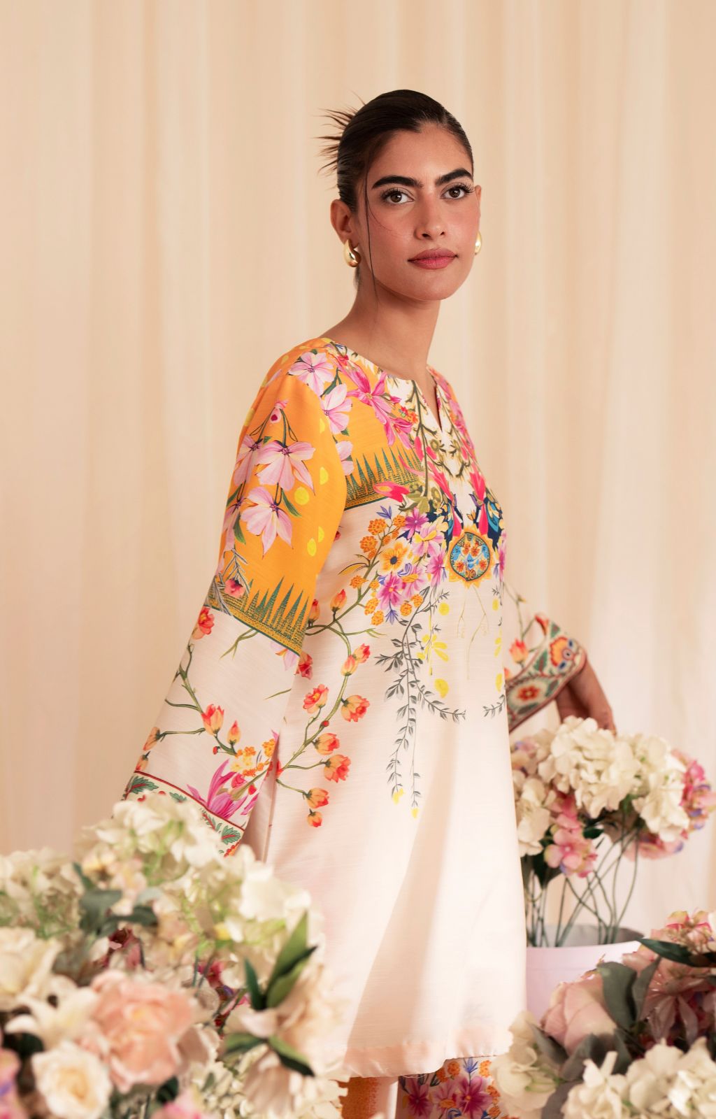 Woman wearing a floral dress with a yellow sleeve, standing among flowers against a beige curtain.
