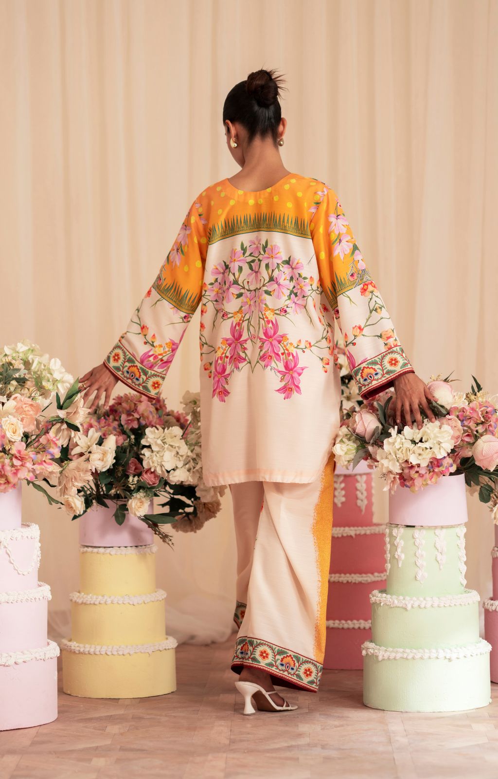 Woman in a colorful traditional outfit standing among floral arrangements on a wooden floor.