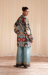 Woman wearing a patterned top and light blue pants against a beige curtain background